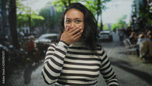 Foto Woman hand on chest and hand to mouth on street in striped top, open mouth and raised eyebrow surprised expression; surprise wonder