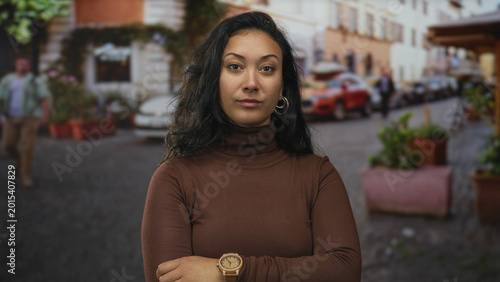 Quadro em tela Woman with hand on chin and hoop earring on a cobblestone street beside potted planters and parked cars wearing brown turtleneck; thoughtful confidence