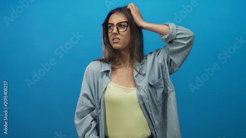 Foto Woman with glasses and hand on head, looking sideways with a grimace, wearing denim shirt and yellow camisole against bright blue background in studio; frustration