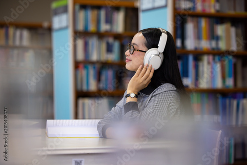 Young Asian woman smiles while listening to music in library. She wears glasses and headphones, Bookshelves line both sides of the aisle behind her. Soft light illuminates her focused. Education