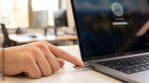 Close-up shot showing a person's hand using a finger to touch a security key (hardware token) plugged into the side of a laptop. The screen displays a login interface