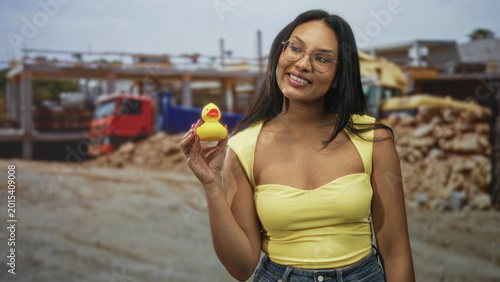 Billede på lærred Woman holding rubber duck up while smiling, bare shoulder visible, wearing yellow tank top and jeans at building; playful nostalgia