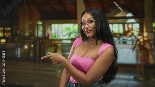 Foto Woman points finger to the right showing index finger and pink top in a building interior with glasses and denim; playful confidence