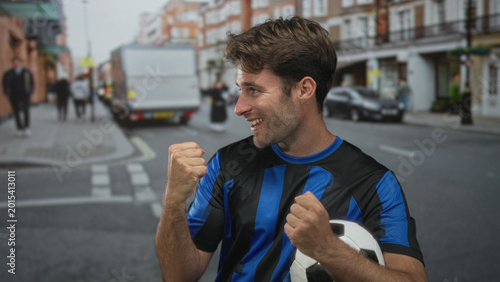Slika na platnu Man clenches fists and holds soccer ball on a city street with cars and pedestrians nearby, smiling broadly; team pride joy