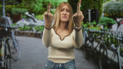 Foto Young redhead woman shows both middle fingers with hands extended toward camera on a street lined with bicycles and iron railing; defiance rebellion attitude