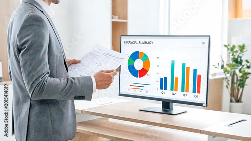 Businessman in Gray Suit Standing at Office Desk Reviewing Financial Dashboard Report Documents with Pie Chart and Bar Chart on Computer Monitor for Business Financial Summary Analysis