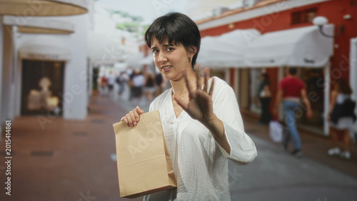 Foto Woman holding paper bag with palm out stop gesture on a busy street, grimacing as if smelling something unpleasant; disgust refusal