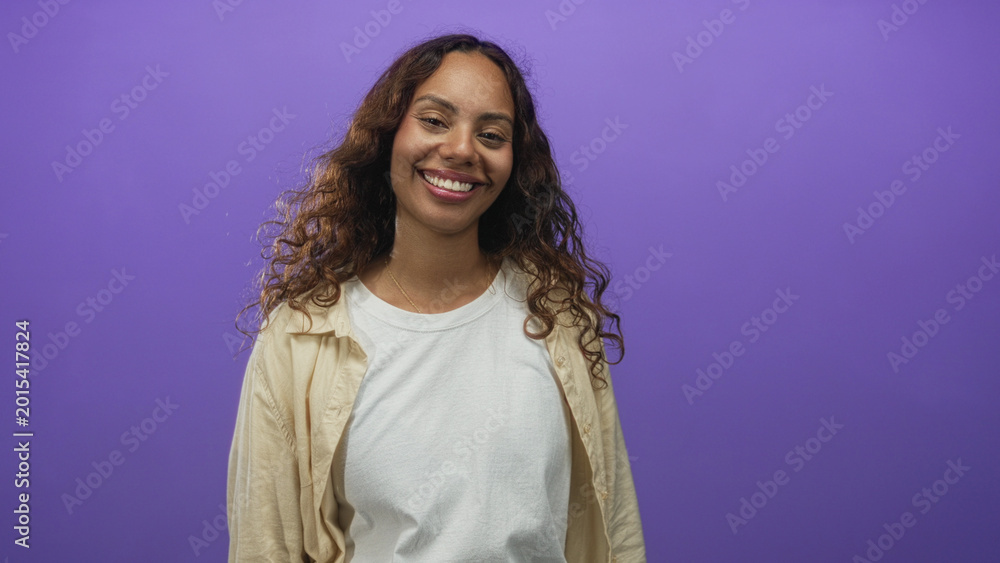 Fototapeta premium Woman smiling with eyes closed showing teeth and loose curly hair, wearing white t shirt and beige overshirt in a purple studio; contentment.