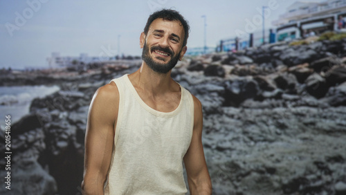 Photos Man smiling with bare arms and beard, wearing sleeveless top posed before a rocky seafront backdrop in a studio; contentment seaside nostalgia