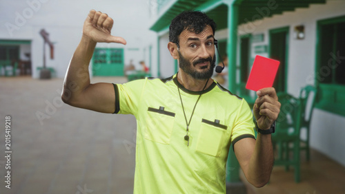 Photos Man referee with beard holding a red card and whistle showing thumbs down gesture in a building corridor; discipline disapproval