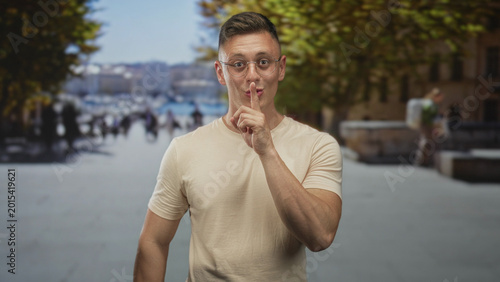 Fotografie Man wearing glasses, finger to lips for silence on pedestrian street by waterfront and trees and benches; quiet secrecy