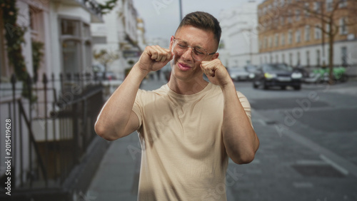 Foto Man posing with fists near his face and smiling on a street in front of parked vehicles and townhouses, wearing glasses and a beige t shirt; playful confidence