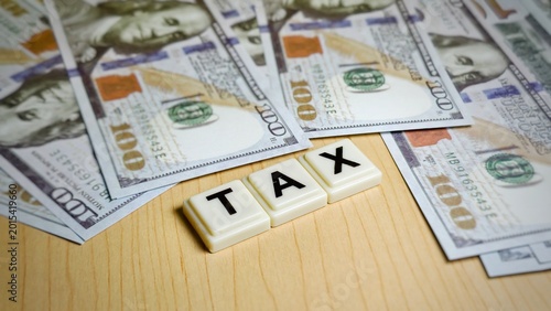 Bild auf Leinwand "TAX" are positioned on a light wooden surface surrounded by various US $100 banknotes, representing government levies, fiscal policy, and mandatory financial contributions