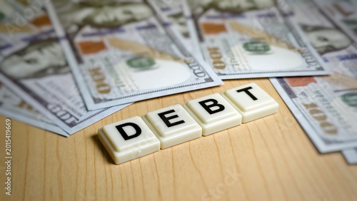 Schilderij op canvas The word DEBT is spelled with tile blocks on a wood surface, surrounded by scattered US $100 bills, illustrating financial obligation, borrowing, and the concept of monetary deficit