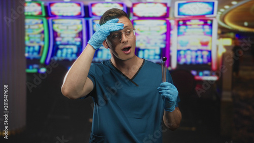 Photography Man dentist in blue scrubs and gloves holds dental mirror, gloved hand to forehead in a casino building; surprise curiosity