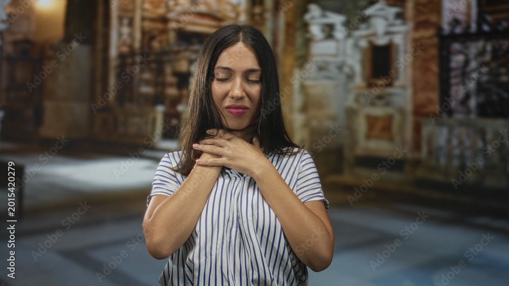 Fototapeta premium Hispanic woman with hands on throat inside a church building, eyes closed in apparent choking pose; panic plea.