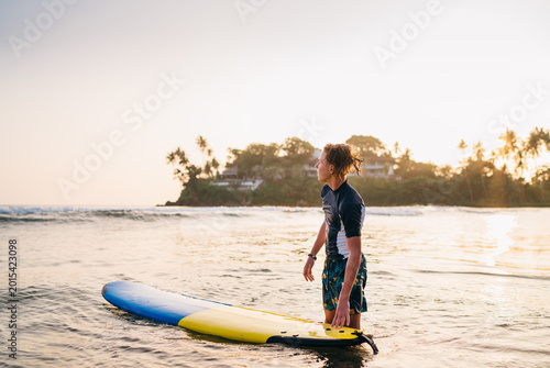 Young teenager boy with colorful surfboard go into sea for surfing. He enjoying a beautiful sunset light on Dewata beach on Sri Lanka. Active sport vacation and exotic countries traveling concept