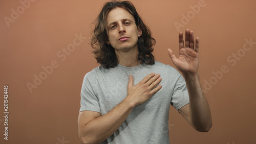 Fotografi Man with long hair placing hand on chest in peach studio, bare forearm visible; sincerity reflection