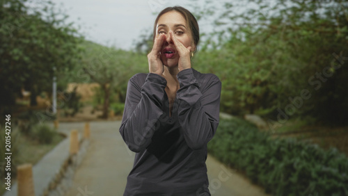 Fotografia Woman cupping hands around mouth shouting on a street path lined with shrubs and wooden bollards; urgency warning