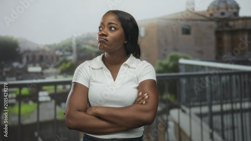 Canvas Print Woman with crossed arms on a building balcony overlooking historic ruins, pouting and looking sideways with tense posture; annoyance