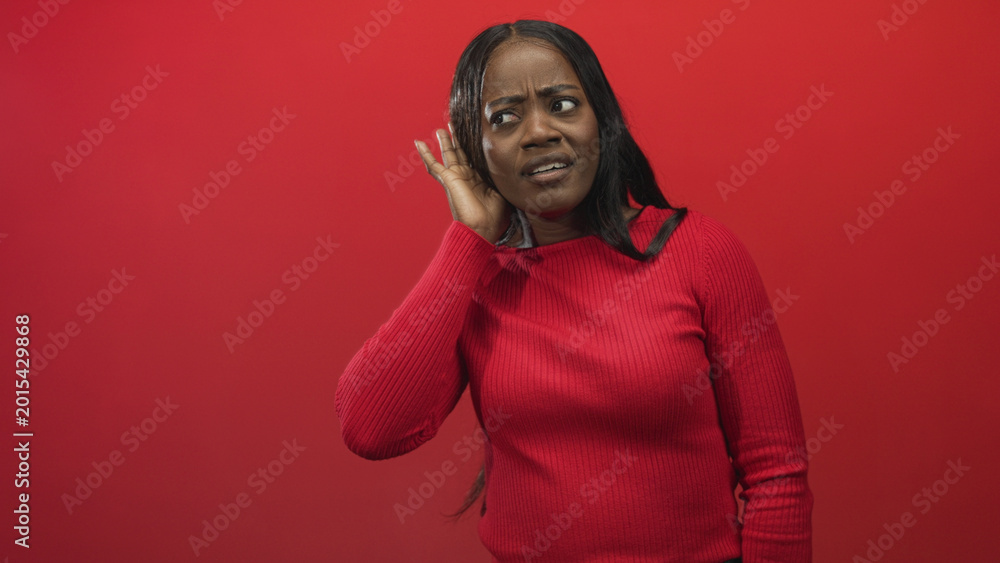 Obraz premium Woman cupping ear with hand listening carefully in studio red backdrop wearing ribbed red sweater; curiosity playfulness.