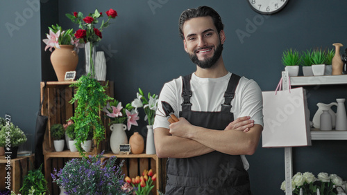 Quadro em tela Man florist holding pruners with crossed arms among vases and flowers in building shop interior, smiling at camera; friendly confidence