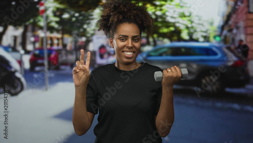 Cuadro en lienzo Young african american woman holding a dumbbell and making a peace sign on a busy city street while smiling to camera; confidence wellness