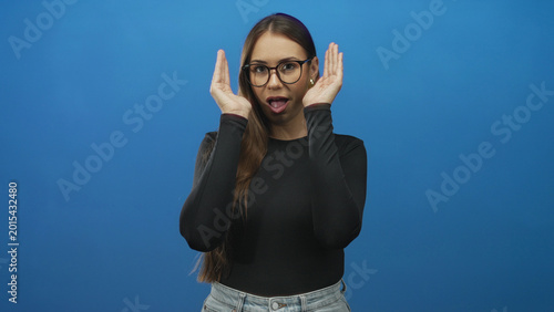 Bild auf Leinwand Woman with glasses lifts both hands to frame her face and briefly covers her eyes while pouting in a blue studio; playful curiosity