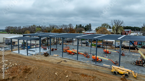 Workers are building a large structure using steel beams and machinery on a construction site. Various tools and equipment are visible along with nearby buildings and trees.