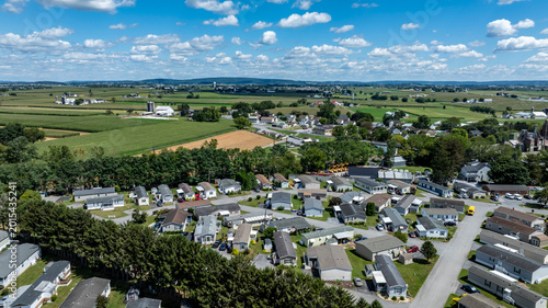 Houses are arranged in a neighborhood surrounded by fields and trees. Bright sky with clouds adds to the scene. This shows a typical residential setup in a rural area.