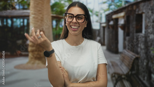 Fotografi Young brunette woman wearing oversized glasses and finger pointing while smiling at camera on a tree lined street with bench; playful banter