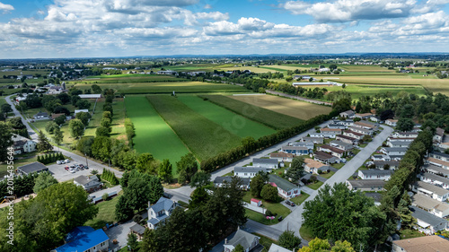A view of a small town surrounded by green fields and farm areas. The houses are in neat rows, while the fields stretch across the landscape. The sky is partly cloudy and blue.
