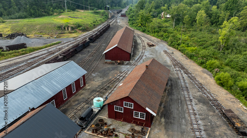 The scene shows multiple red storage buildings near train tracks. The area is green with trees and a slight incline in the background. Equipment and materials are scattered around the site.
