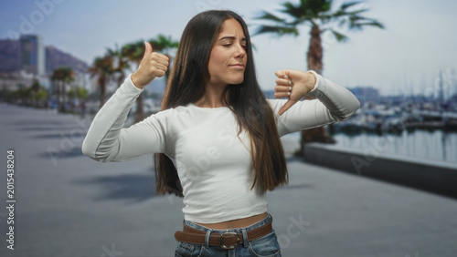 Fotografie Woman showing thumbs up and thumbs down on a street near a marina and palm trees; ambivalence decision