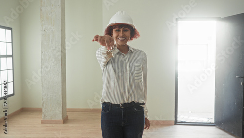 Fotografie Woman holding keys with outstretched hand in an empty building room, smiling while wearing hardhat and striped shirt as architect presenting access; homeowner pride