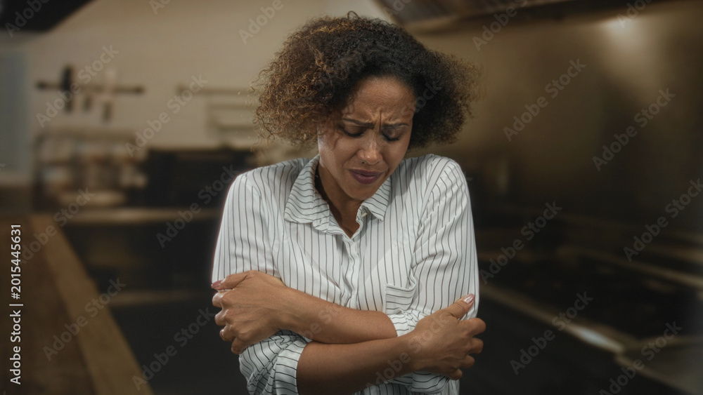Fototapeta premium Woman hugging her arms across chest in a kitchen building, eyes closed and wincing; discomfort tension.