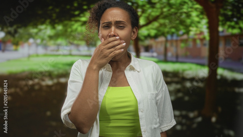 Fotografie Woman with hand covering mouth and arms outstretched in forest wearing green crop and white shirt, midriff visible; fatigue
