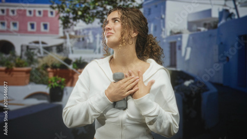 Canvas Print Woman holding a dumbbell to her chest, hands clasped over heart on a sunlit street courtyard with mediterranean buildings; recovery relief