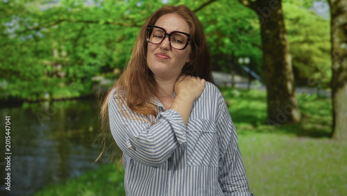 Canvas Print Young caucasian bespectacled woman in striped shirt holds her sore shoulder beside tree in grassy forest clearing; physical discomfort