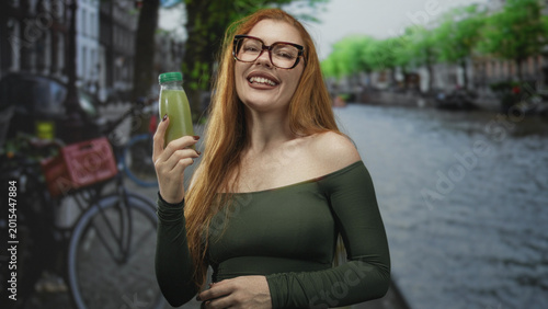 Photography Redhead woman holding green juice bottle, bare shoulder exposed, smiling on street by a canal; carefree joy