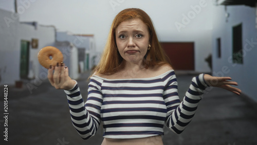 Cuadro en lienzo Young redhead woman in striped off shoulder top holds a doughnut in left hand, pouts and shrugs right hand in a narrow white street; indecision