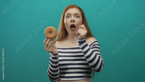 фотографія Young redhead woman holding a glazed donut, finger to chin thinking with skeptical expression and bare shoulder visible in studio; doubt