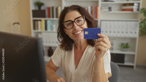 Cuadro en lienzo Woman holding blue creditcard with hand, smiling at monitor while seated at desk in building; confidence convenience