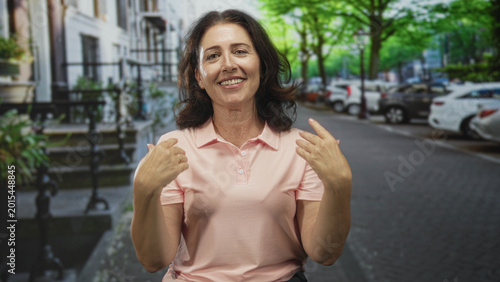Fotografie Woman in pink polo pointing both hands toward camera on a tree lined street with parked cars and stoop steps; invitation friendly