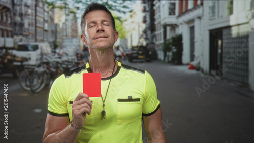 Foto Referee man in fluorescent yellow shirt holding a red card and whistle at chest, eyes closed, standing on a city street with parked bikes; authority
