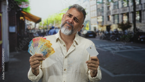 Tableau sur toile Man holding fanned swiss franc banknotes and white card on street in city, hands visible and card shown; financial uncertainty