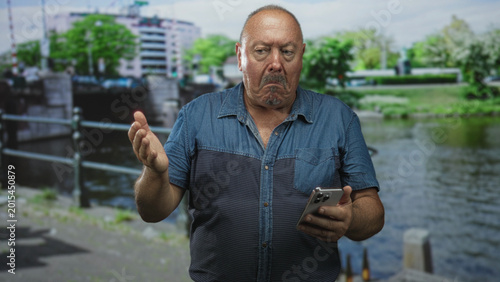 Billede på lærred Man holding smartphone with palm up gesture and furrowed brow on street riverside pavement; frustration concern