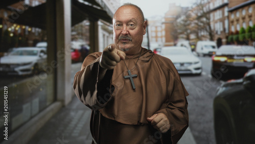 Tableau sur toile Senior monk man points finger wearing cross necklace on city street with cars and storefronts; faith authority