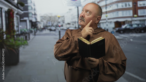 Obraz na plátne Man monk holds bible, finger to chin in a thinking gesture on street with robe visible; serenity contemplation