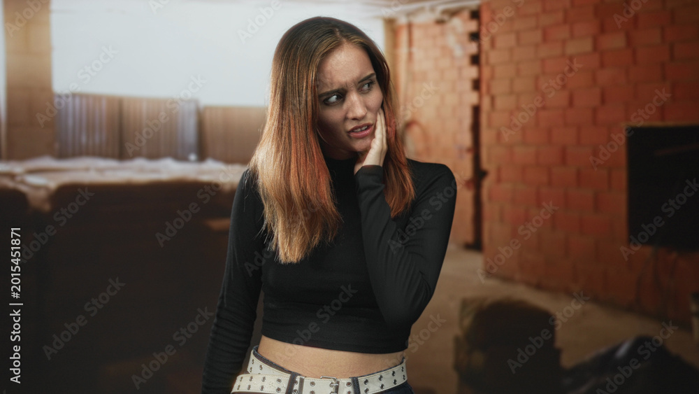 Fototapeta premium Woman hand to cheek with grimace, touching jaw and glancing sideways in unfinished brick building with mattress and window light; anxiety unease isolation.